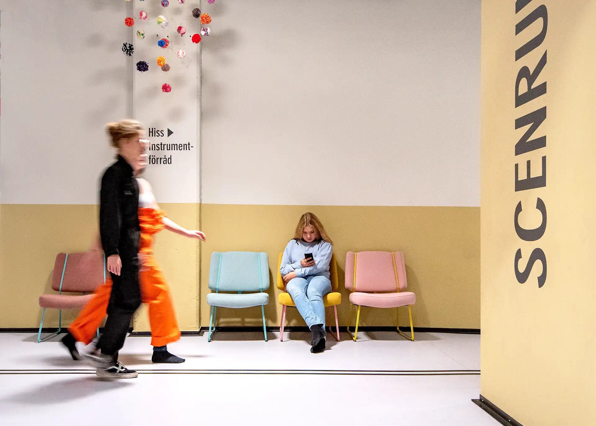 Foto på två elever som rör sig i en korridor. En elev sitter på en av de tre fåtöljerna som syns på bilden. Hen tittar ned på sin mobiltelefon.